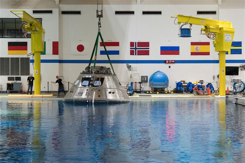 A space capsule is suspended from the ceiling. It rests on the surface of water in a pool.