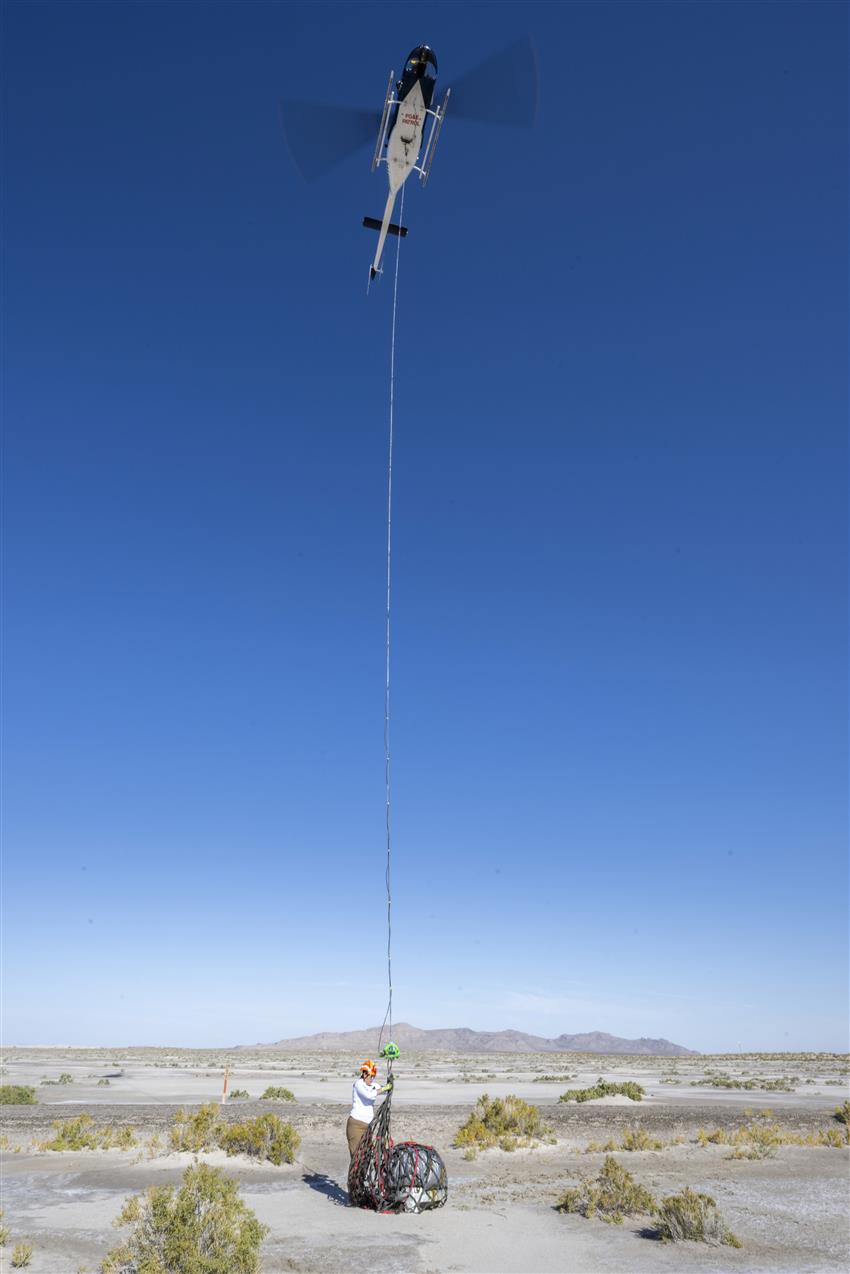 Dans un désert, quelqu’un attache un filet où se trouve une capsule spatiale à un câble descendu d’un hélicoptère.
