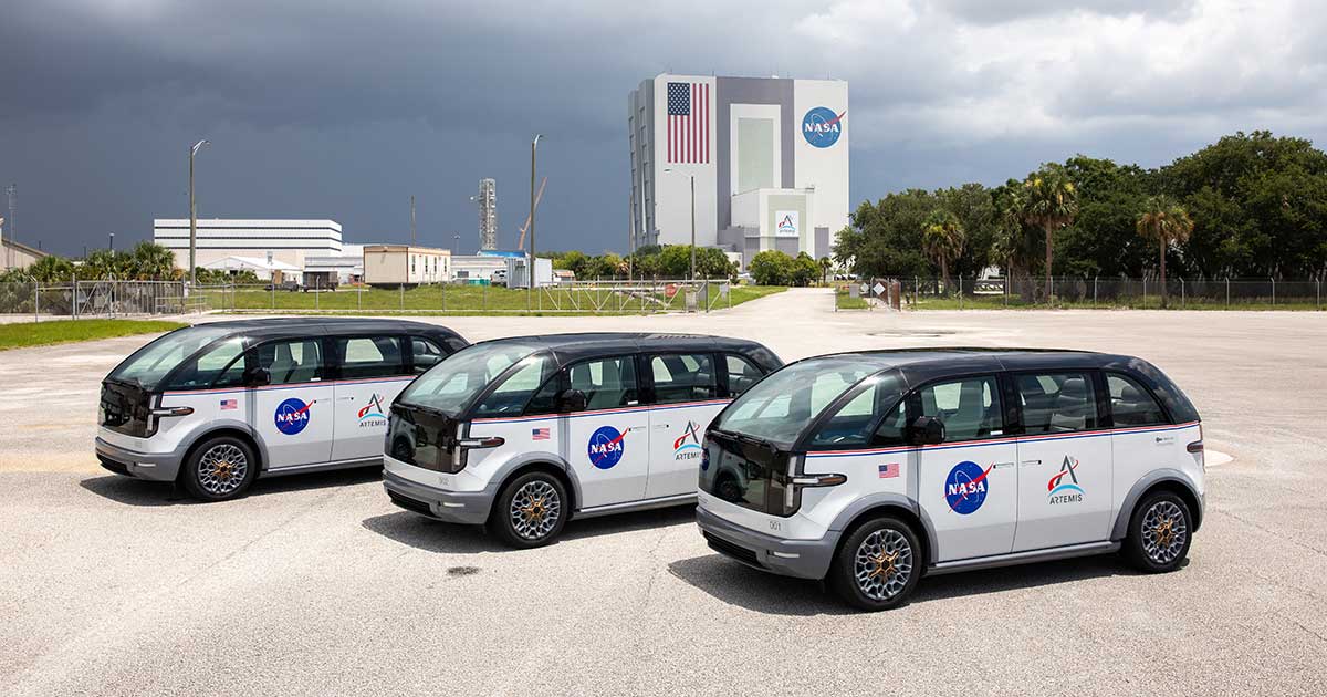 Three van-sized vehicles are parked in a row. NASA's Vehicle Assembly Building is prominent in the background.