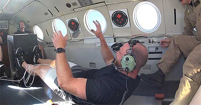 A man exercises using a flywheel aboard an airplane. Objects float around him.