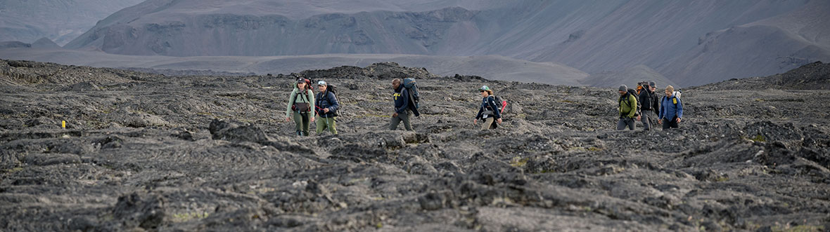 Sept personnes portant des sacs à dos se déplacent sur un terrain rocheux. Il y a de grandes montagnes désertiques à l'arrière-plan.