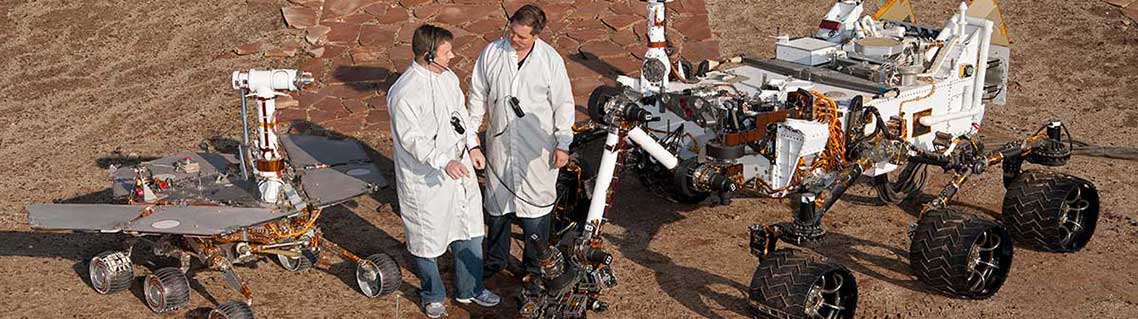 Two spacecraft engineers stand with three generations of Mars rovers.