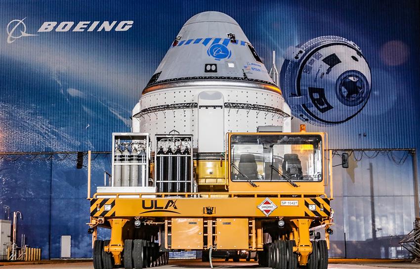 Un vaisseau spatial sur une plateforme de transport dans le hall d’intégration de Boeing, où une murale montre le logo.
