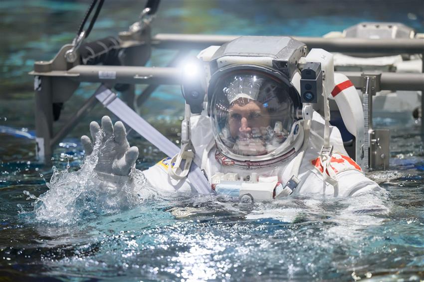 Joshua is wearing a spacesuit while being lowered into a pool.