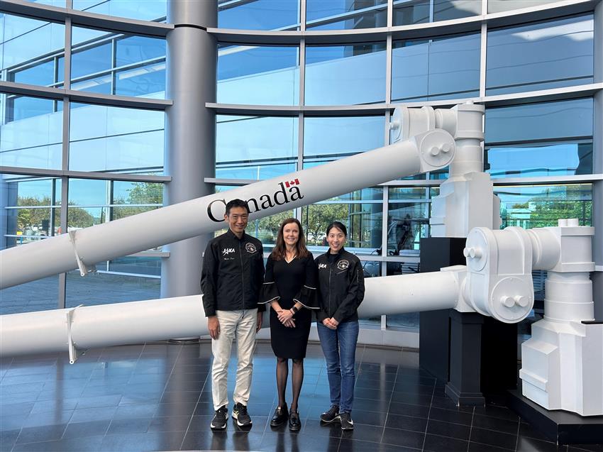 Makoto Suwa, Lisa Campbell and Ayu Yoneda standing in front of a replica of the Canadarm2 robotic arm.