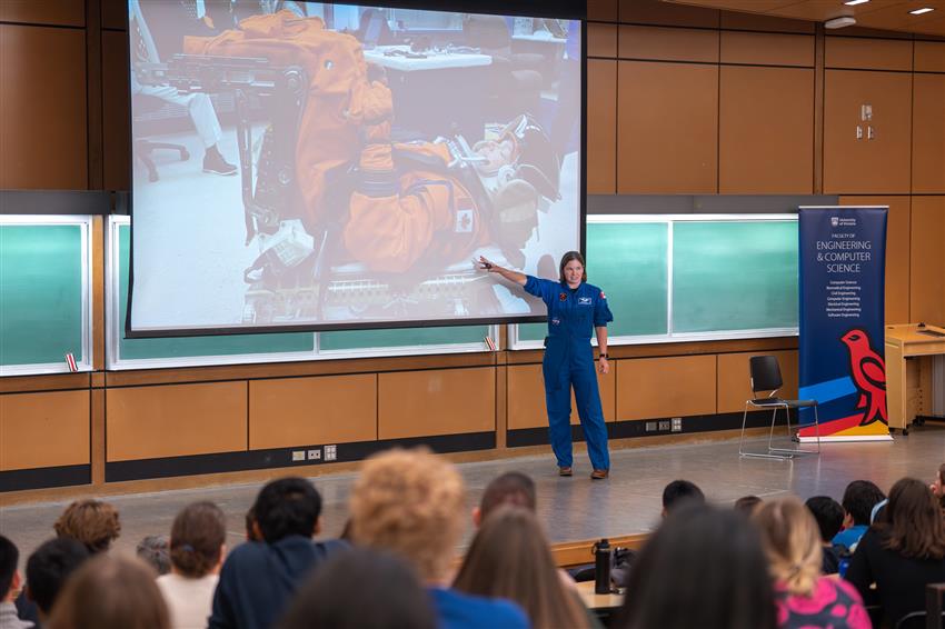 Jenni, in a university auditorium, points to a large screen with a photo of her in an orange spacesuit.