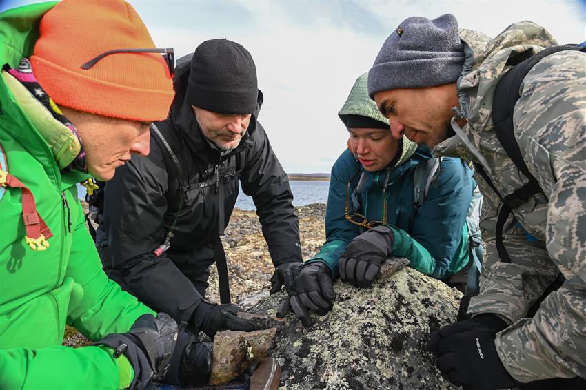 Quatre scientifiques accroupis autour d'un gros rocher. On aperçoit un plan d'eau derrière eux.