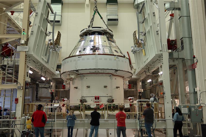 A crane lowers a spacecraft crew module into a building.