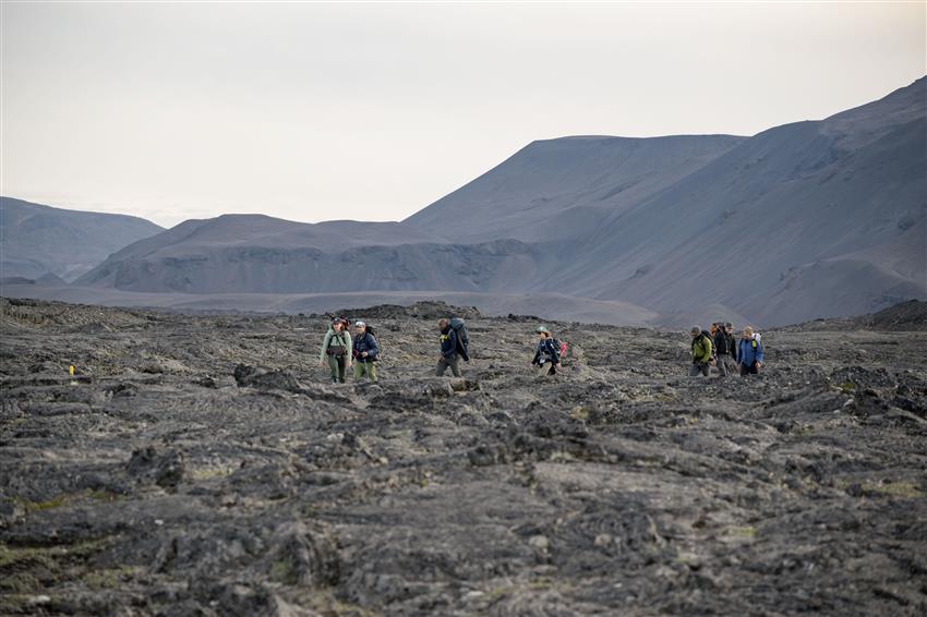 Seven people in warm clothing walk on rocky terrain. There are mountains in the background.