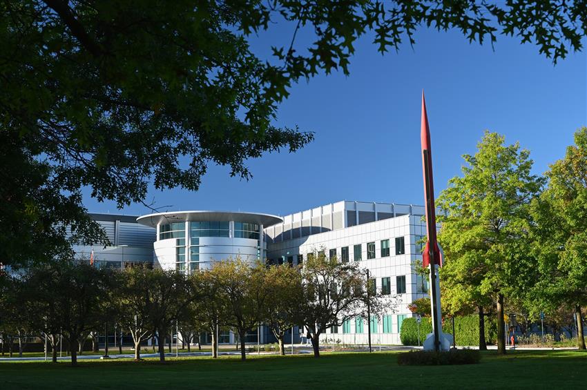 A red and white rocket stands in front of the space centre surrounded by green grass and trees.