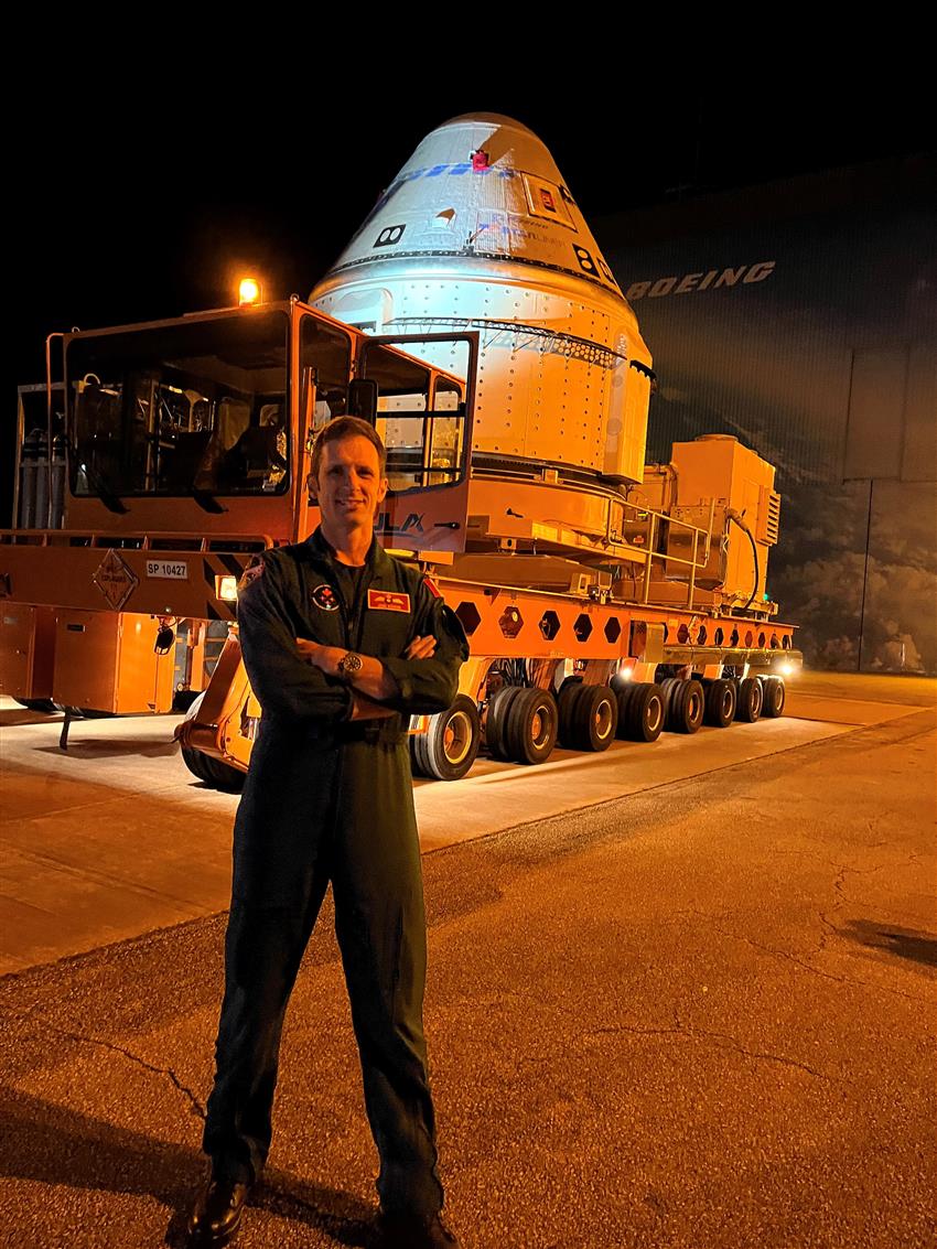 Joshua stands in front of the spacecraft. It is dark out. 