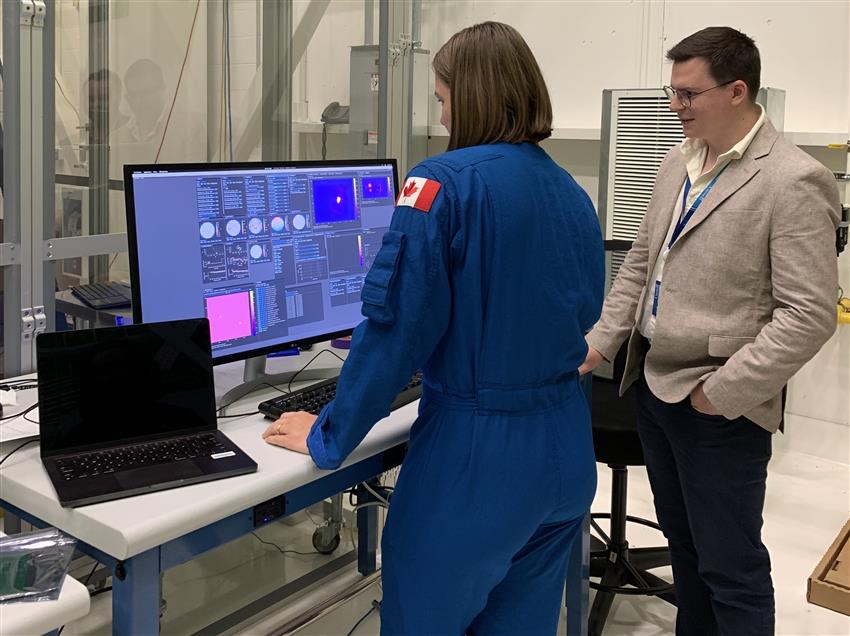 Jenni stands in front of a computer screen with a man to her right.