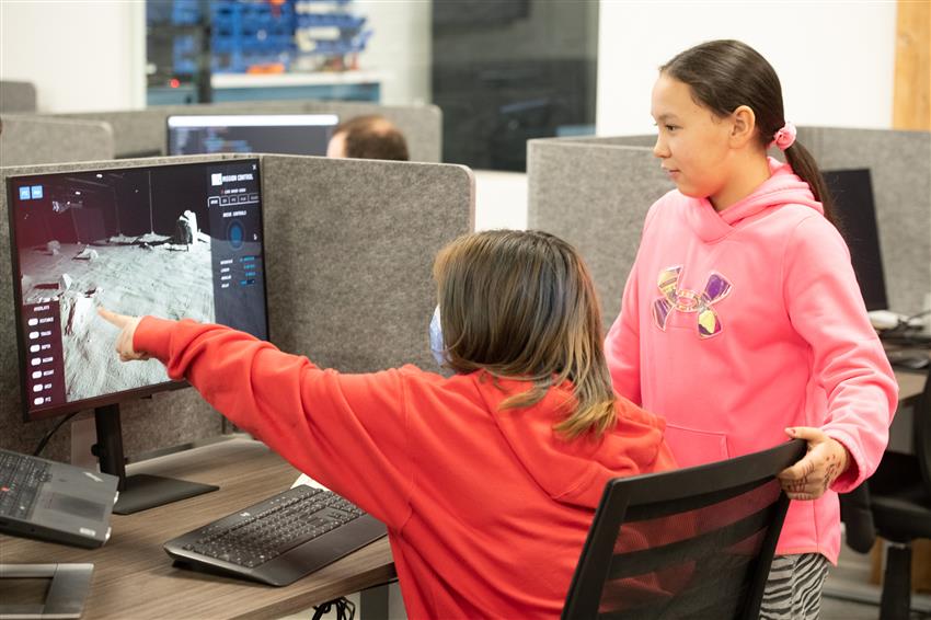 Two girls look at a screen while one of them points to it.