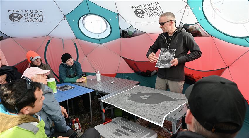 Un homme montre l'image d'un cratère à cinq personnes assises autour d'une table. Ils sont dans une tente.