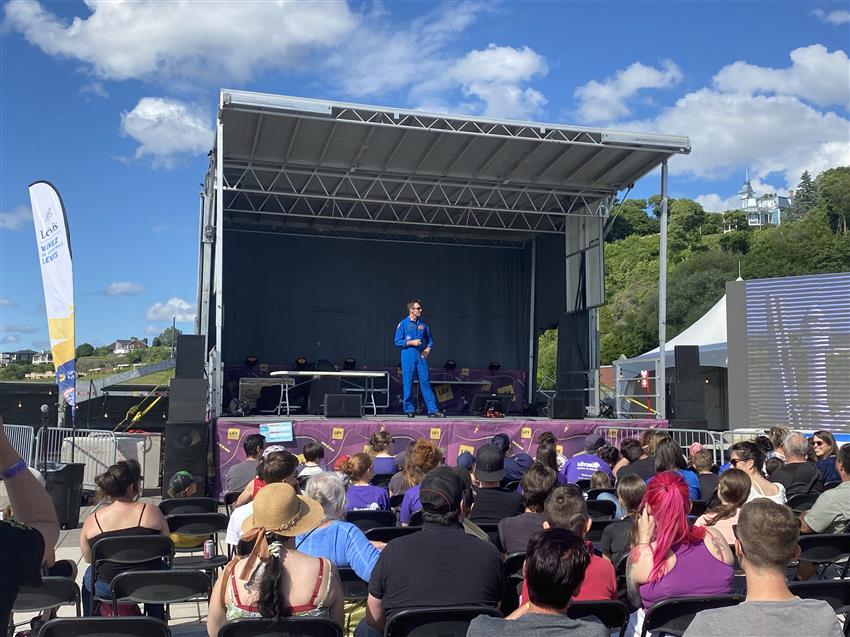 Joshua is standing on an outdoor stage and speaking to the crowd.