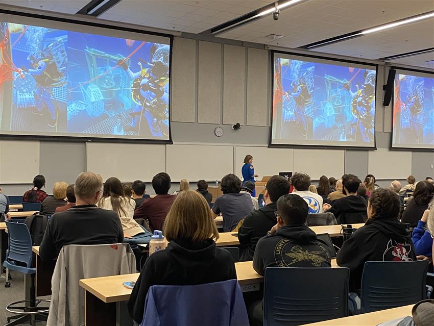 Jenni presents to university students, with three large screens behind her.