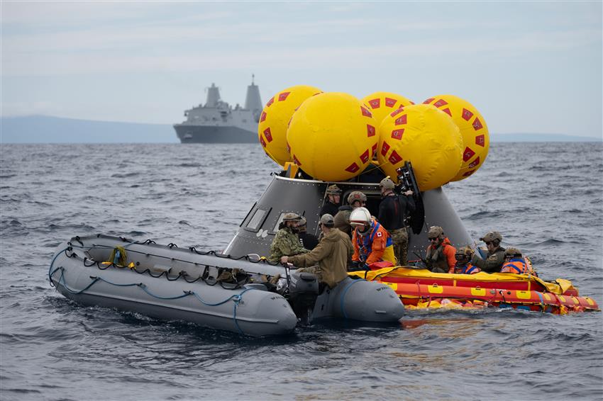 Two inflatable boats carrying people float in front of a cone-size mockup of a spacecraft. 