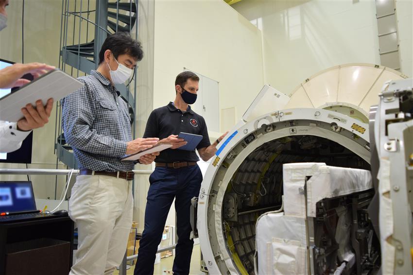 Joshua and Kimiya Yui stand next to the Kibo simulator. They are wearing face masks and holding tablet computers.