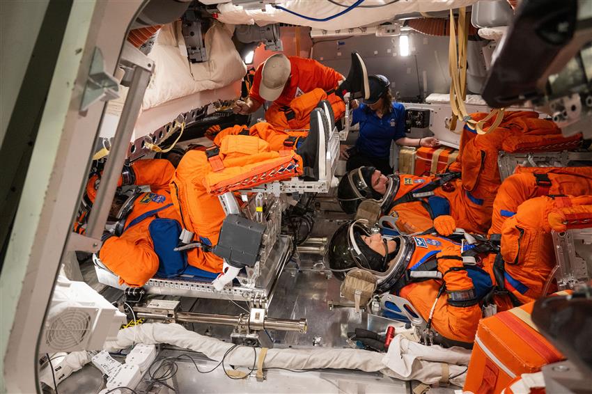 The Artemis II crew in spacesuits sit horizontally with their feet raised in the Orion mockup.