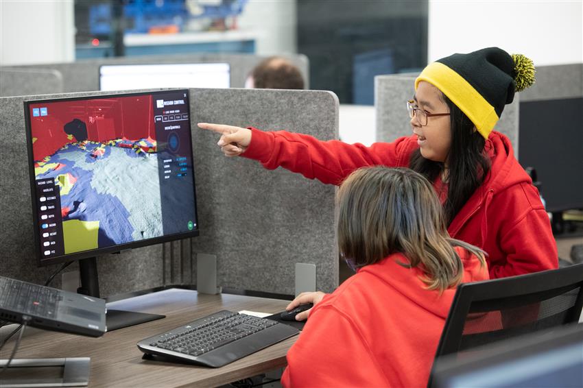 Two girls look at a screen while one of them points to it.