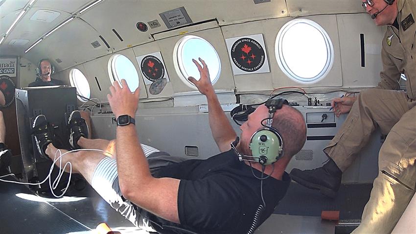 Un homme utilise une roue d'inertie à bord d'un avion. Des objets flottent autour.