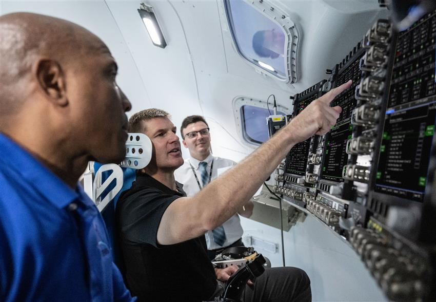 Jeremy is sitting with NASA astronaut Victor Glover and pointing at the screens the Orion simulator.