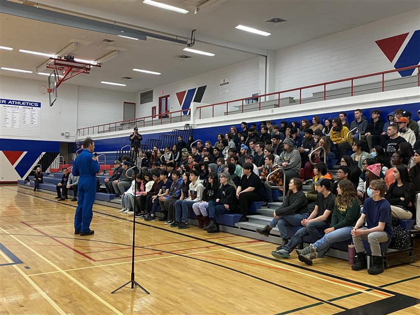 Joshua standing in a gymnasium and speaking to a large group of students.