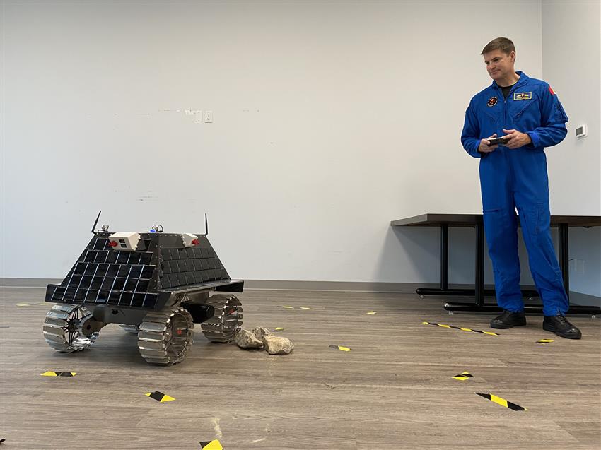 A man in a blue flight suit holds a remote control, a little rover next to him.