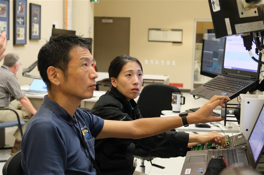 Makoto and Ayu are using different devices in front of a laptop and several screens.