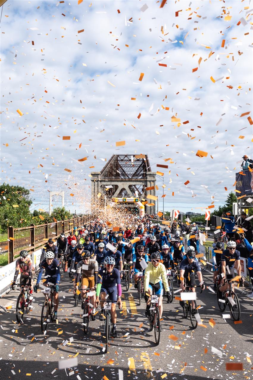 Joshua is surrounded by hundreds of cyclists and confetti with the Quebec bridge in the background.