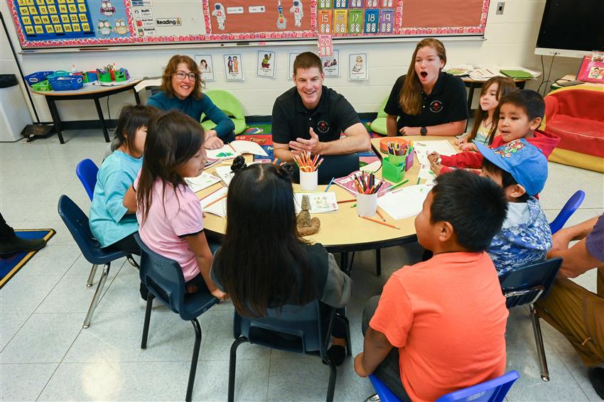Trois adultes et sept enfants sont assis autour d'une table, dans une salle de classe.