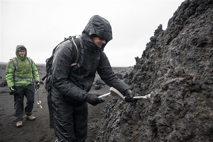 Wearing a wet raincoat, Jeremy uses a hammer and a chisel to break a piece of rock. Reid, in a green coat, stands behind