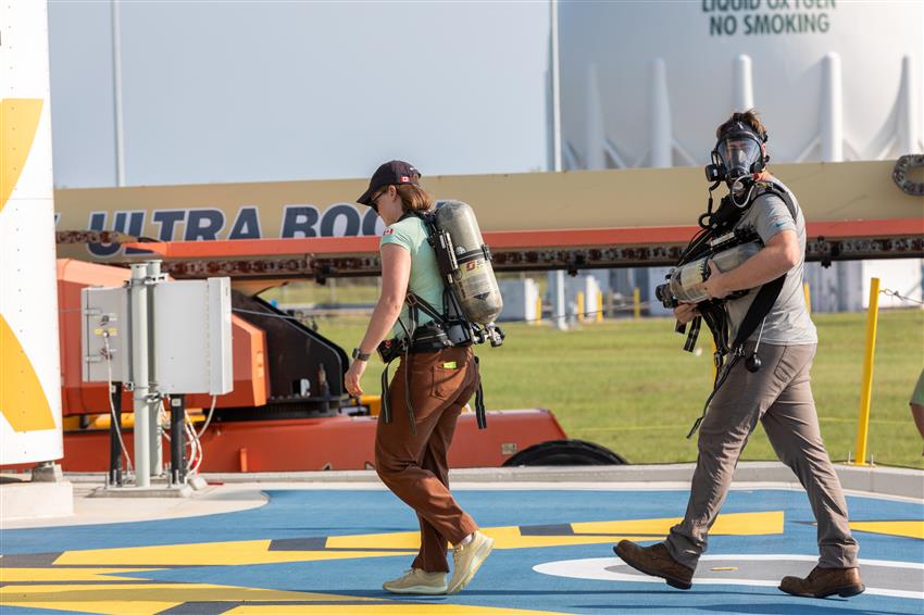 Jenni walks with a man wearing a breathing mask. They both carry oxygen tanks.