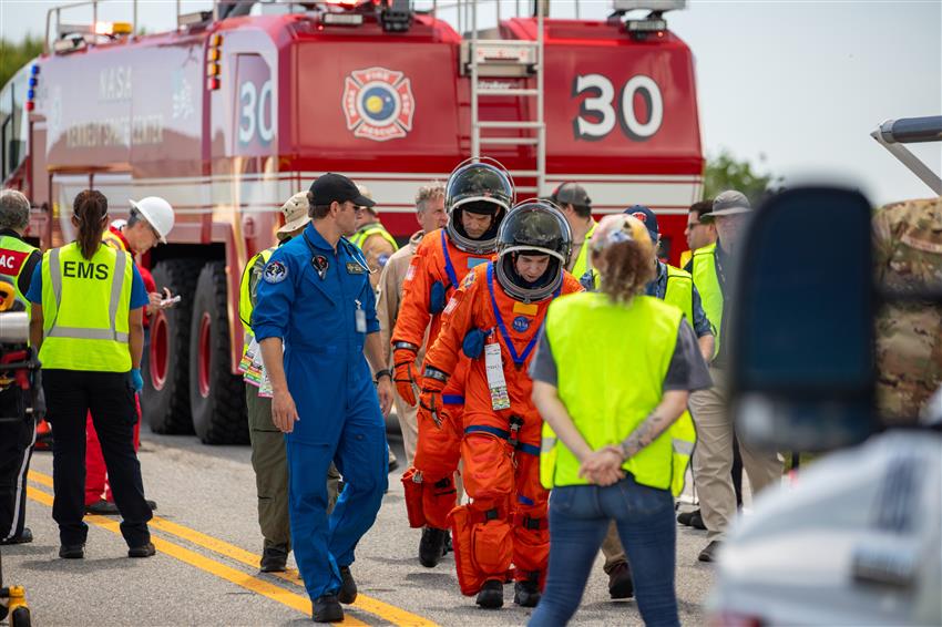 Dr. Greene, in blue, walks with two people in orange suits, an emergency vehicle in the background.