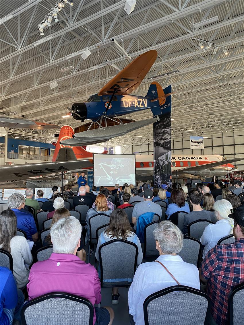 Jeremy speaks to a crowd in a hangar with planes above and behind him.