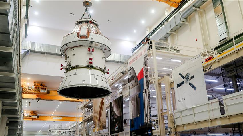 A crane lowers a spacecraft crew module into a building.