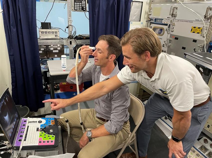 Joshua tient la sonde d'un appareil échographique devant un œil. Le médecin de vol pointe vers un moniteur. 