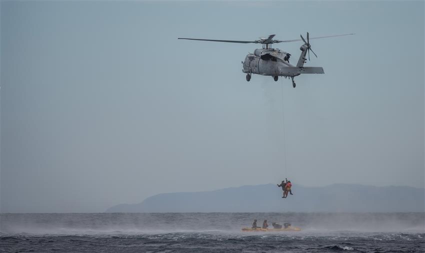 An astronaut is being airlifted by a helicopter, while four other people wait below in an inflatable boat.