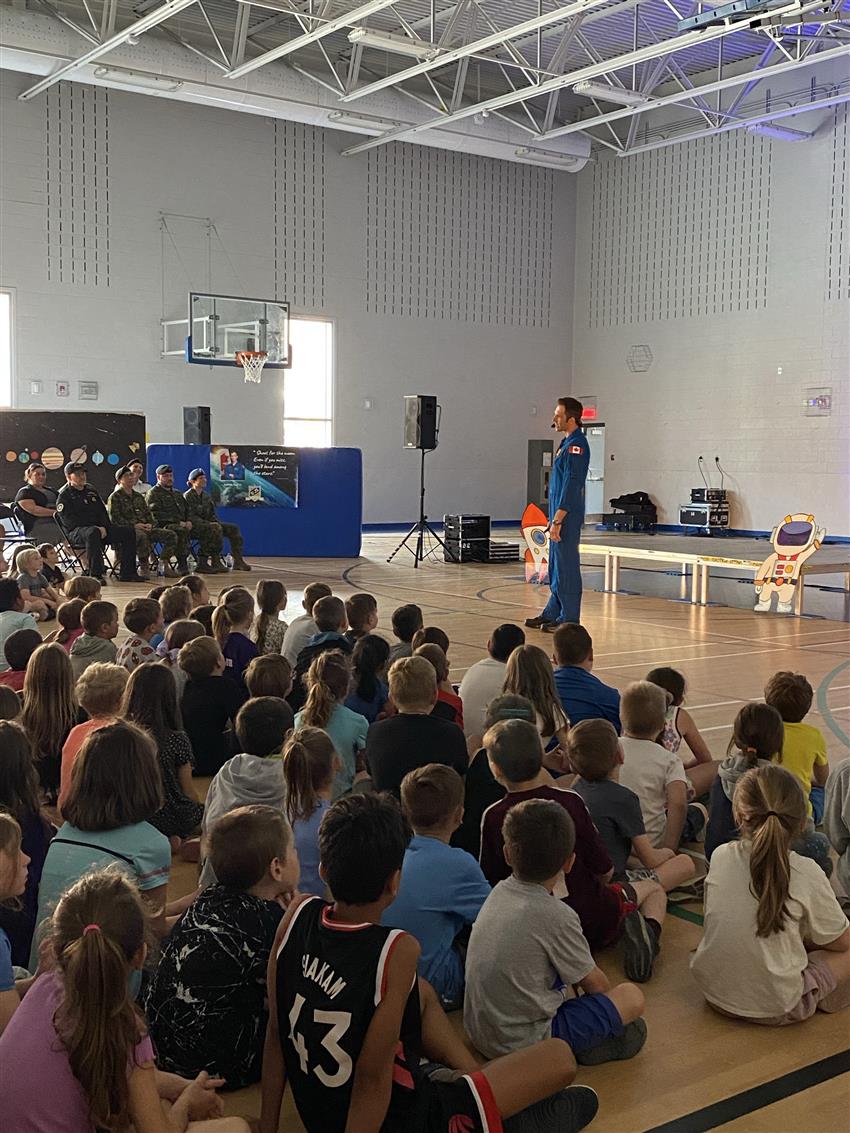 Joshua speaks to an audience of children. Members of the Canadian Forces are sitting to his right.