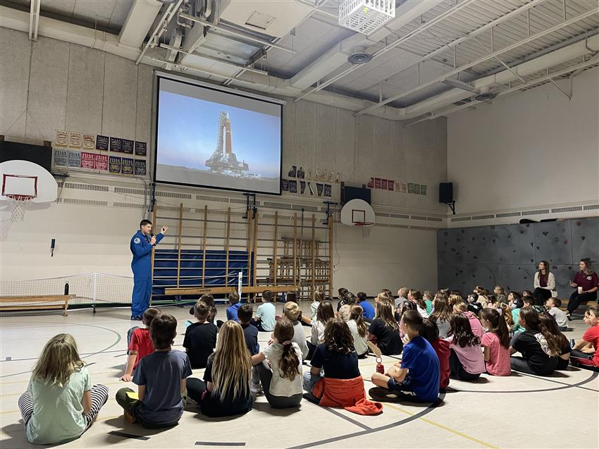 Jeremy speaks to children sitting on a gym floor.