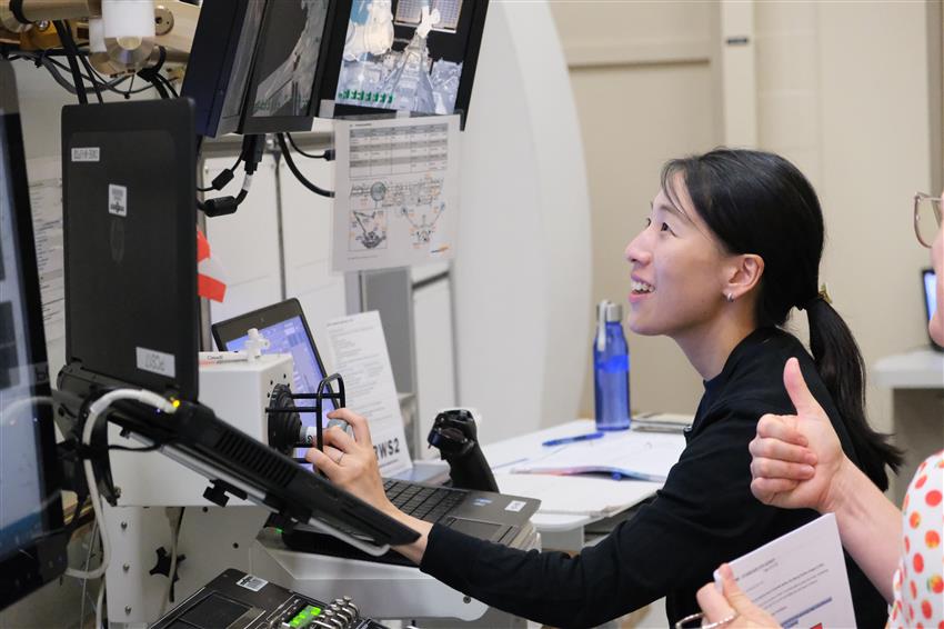 Ayu operates a hand controller in front of a laptop and several screens.