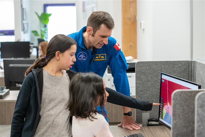 Two girls and Joshua look at a screen. One of them points to it.