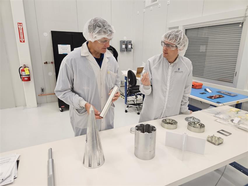 Jeremy and another man wear lab coats and protective gear with metal pieces on a table in front of them.