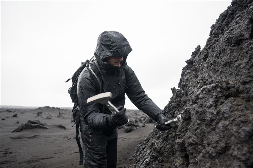 Jeremy is wearing raingear and a backpack. He is about to hit a rockface with a hammer.